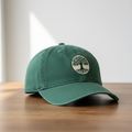 Forest green baseball cap with embroidered tree logo on wooden table with soft natural lighting